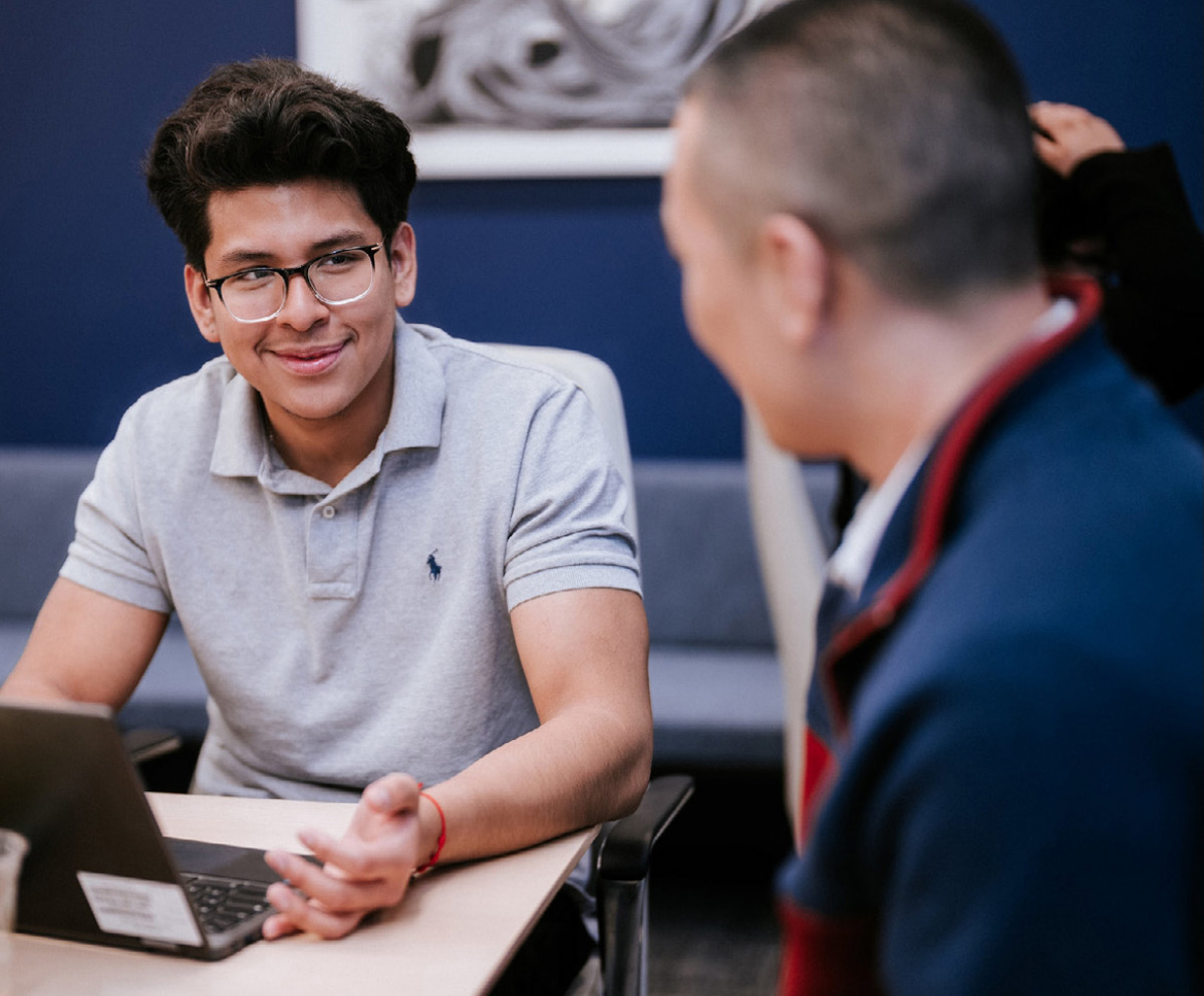 Young man wearing glasses smiling during a mentoring conversation while seated at a table with a laptop.