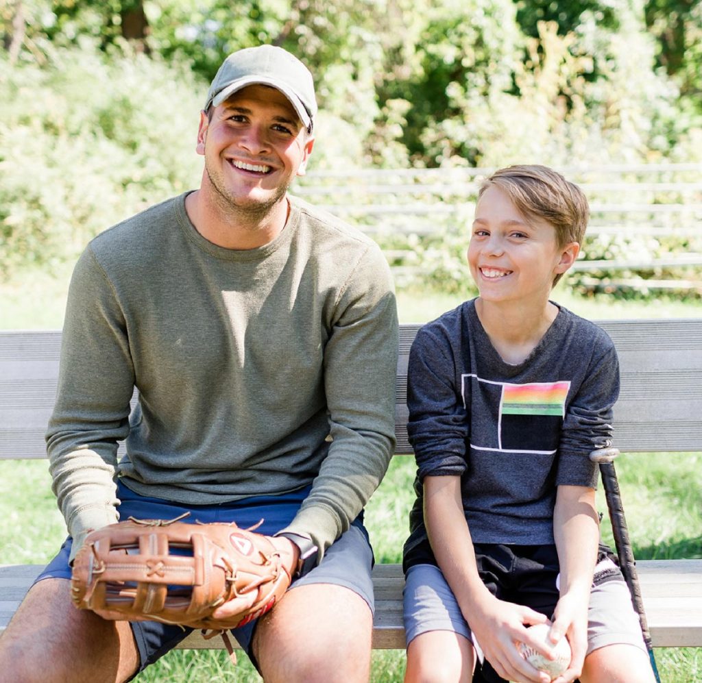 Adult and child sitting on a bench outdoors with baseball gloves and a baseball, smiling together.