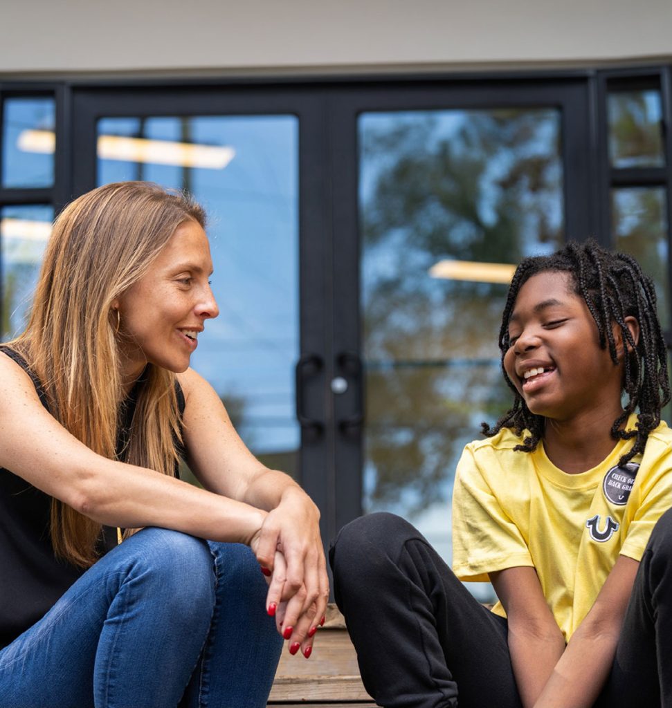 Woman sitting and talking with a smiling child outdoors in front of a building.