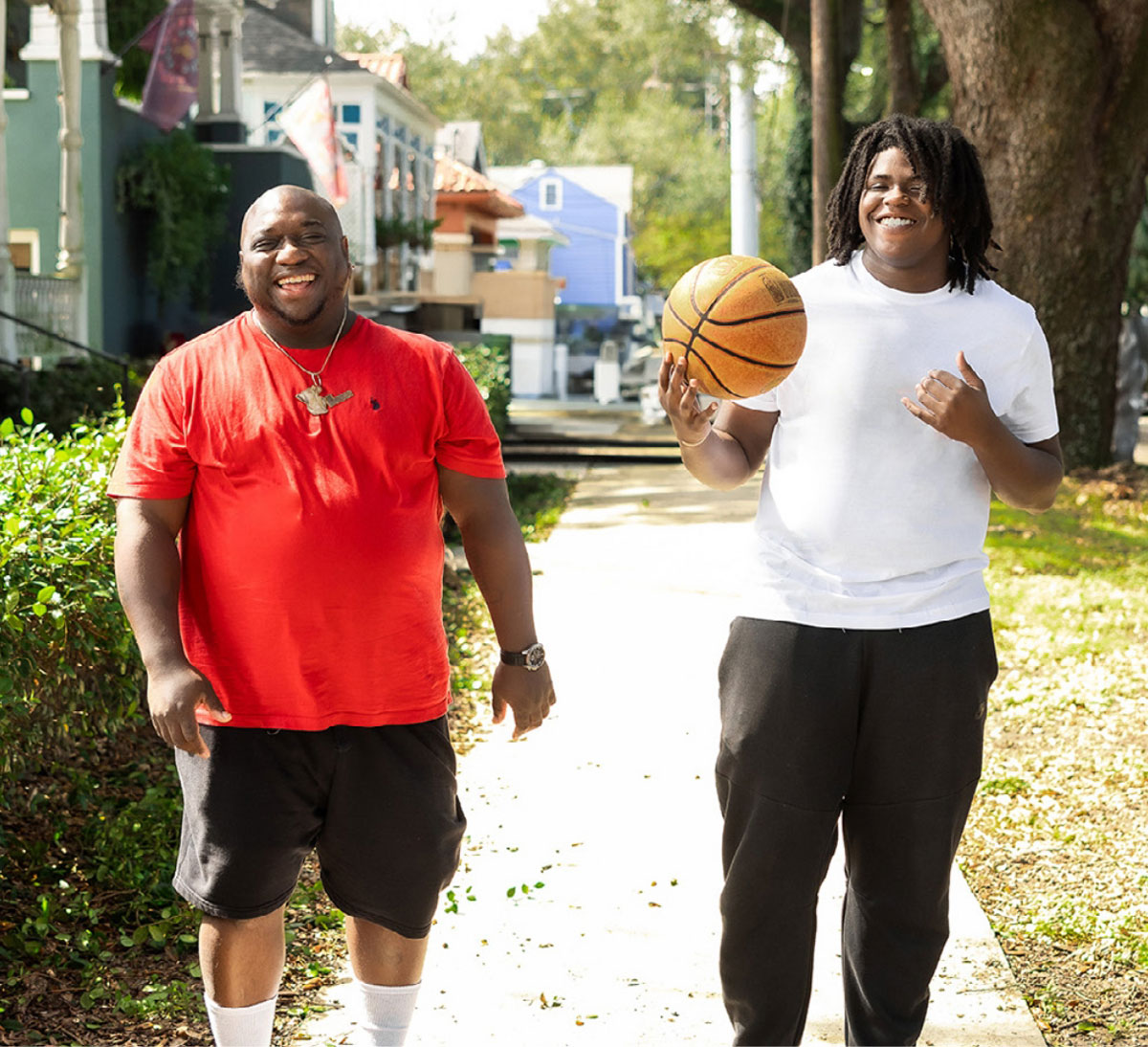 Adult and teenager walking outdoors together while holding a basketball and smiling.
