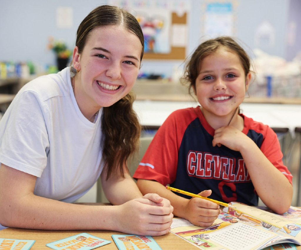 Older student helping a younger child with schoolwork in a classroom setting.